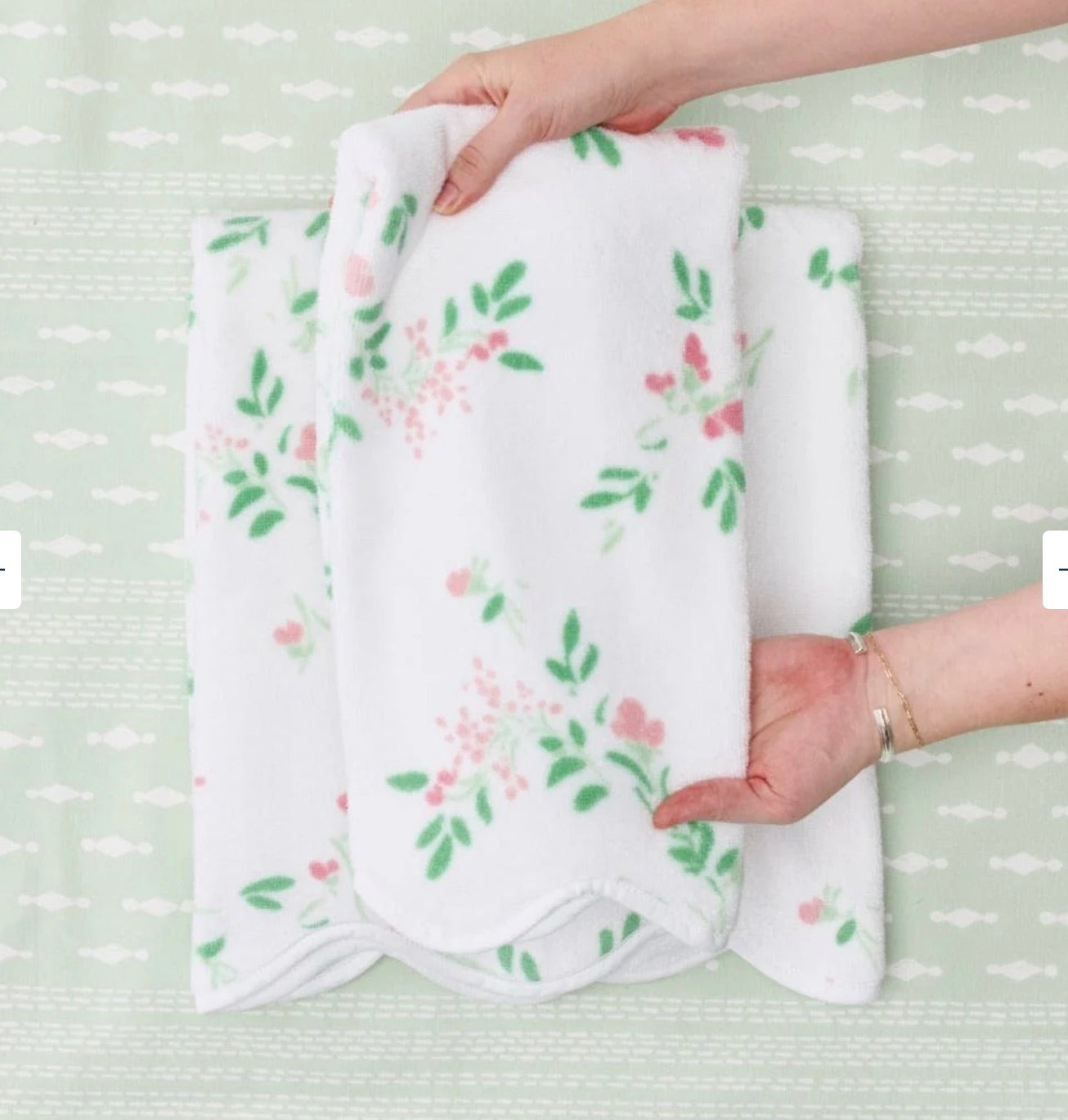 Two hands arrange a white towel from the Alice Floral Scallop Towel Collection by Weezie Towels, featuring a pink and green floral pattern and hand-finished scallop trim, on a light green patterned surface.
