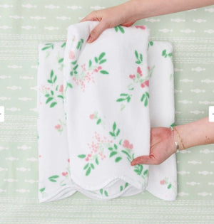 Two hands arrange a white towel from the Alice Floral Scallop Towel Collection by Weezie Towels, featuring a pink and green floral pattern and hand-finished scallop trim, on a light green patterned surface.