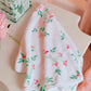 The Alice Floral Scallop Towel from Weezie Towels, featuring a pink and green floral pattern and hand-finished scallop trim, is displayed on a white surface beside a patterned box and pink flower, showcasing its soft, absorbent terry fabric.