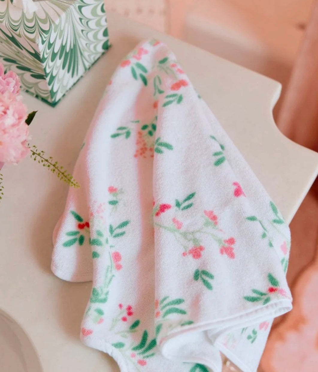 The Alice Floral Scallop Towel from Weezie Towels, featuring a pink and green floral pattern and hand-finished scallop trim, is displayed on a white surface beside a patterned box and pink flower, showcasing its soft, absorbent terry fabric.