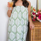A woman in the Ivy Block Towel Wrap by Weezie Towels, featuring a green and white pattern and elastic back, stands hands-free by a wicker table with a vibrant flower arrangement, holding a drink.