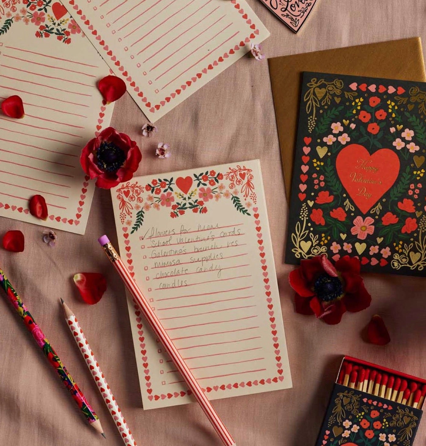 Flat lay of the Rifle Paper Co. Valentine's Day card, Juliet Checklist Notepad, illustrated notepads, pencils, matches, red flower petals, and a handwritten to-do list on a pink surface.