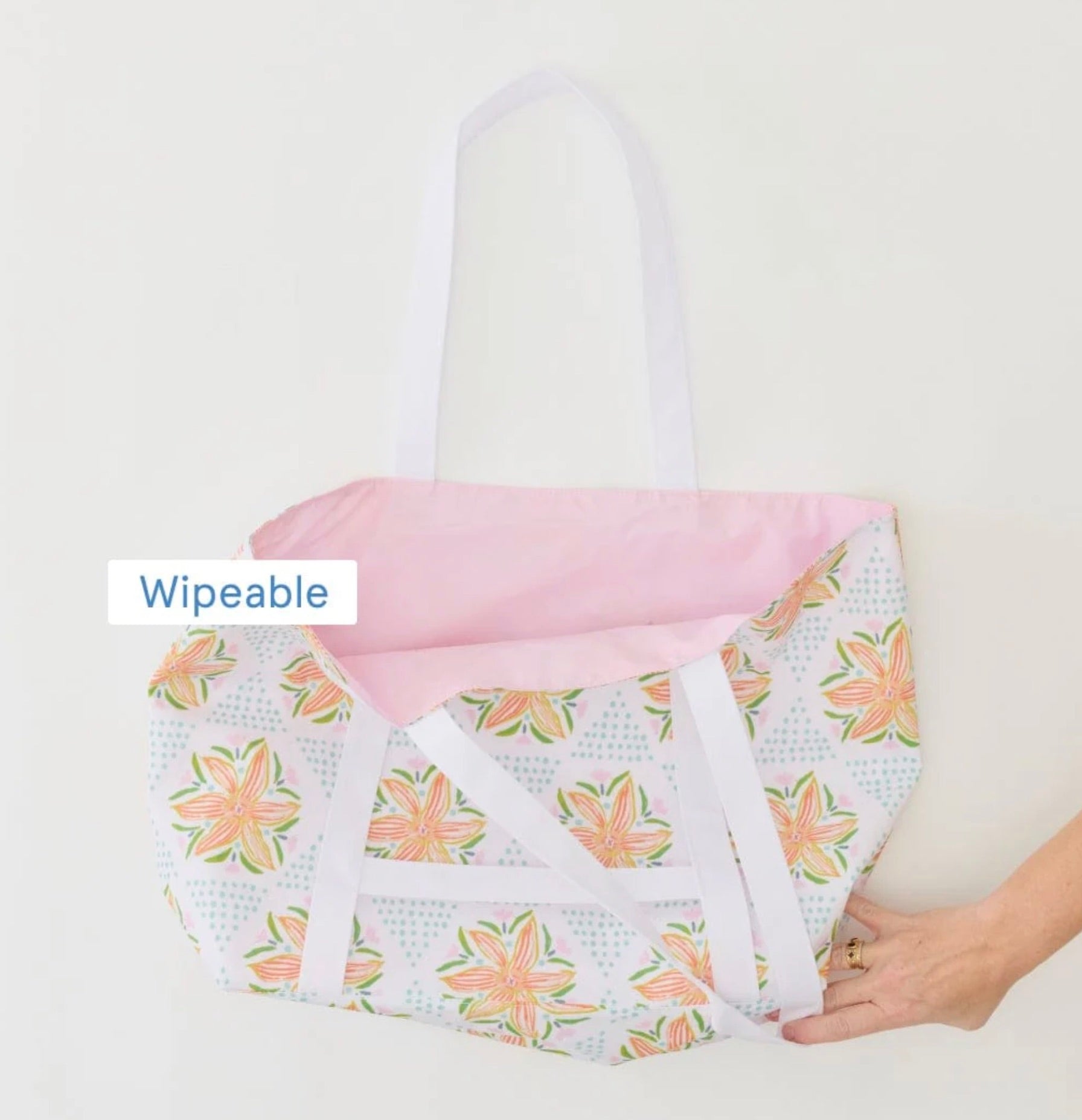 A hand holds the Weezie x Lulie Wallace Beach Tote by Weezie Towels, featuring a Savannah print, white straps, and a pink interior. A "Wipeable" label is displayed on the image.