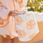 A person in a peach outfit holds the Weezie x Lulie Wallace Wet Bag by Weezie Towels, a floral-patterned, water-resistant bag with a ribbon top—ideal as a wet swimsuit or beach travel bag.
