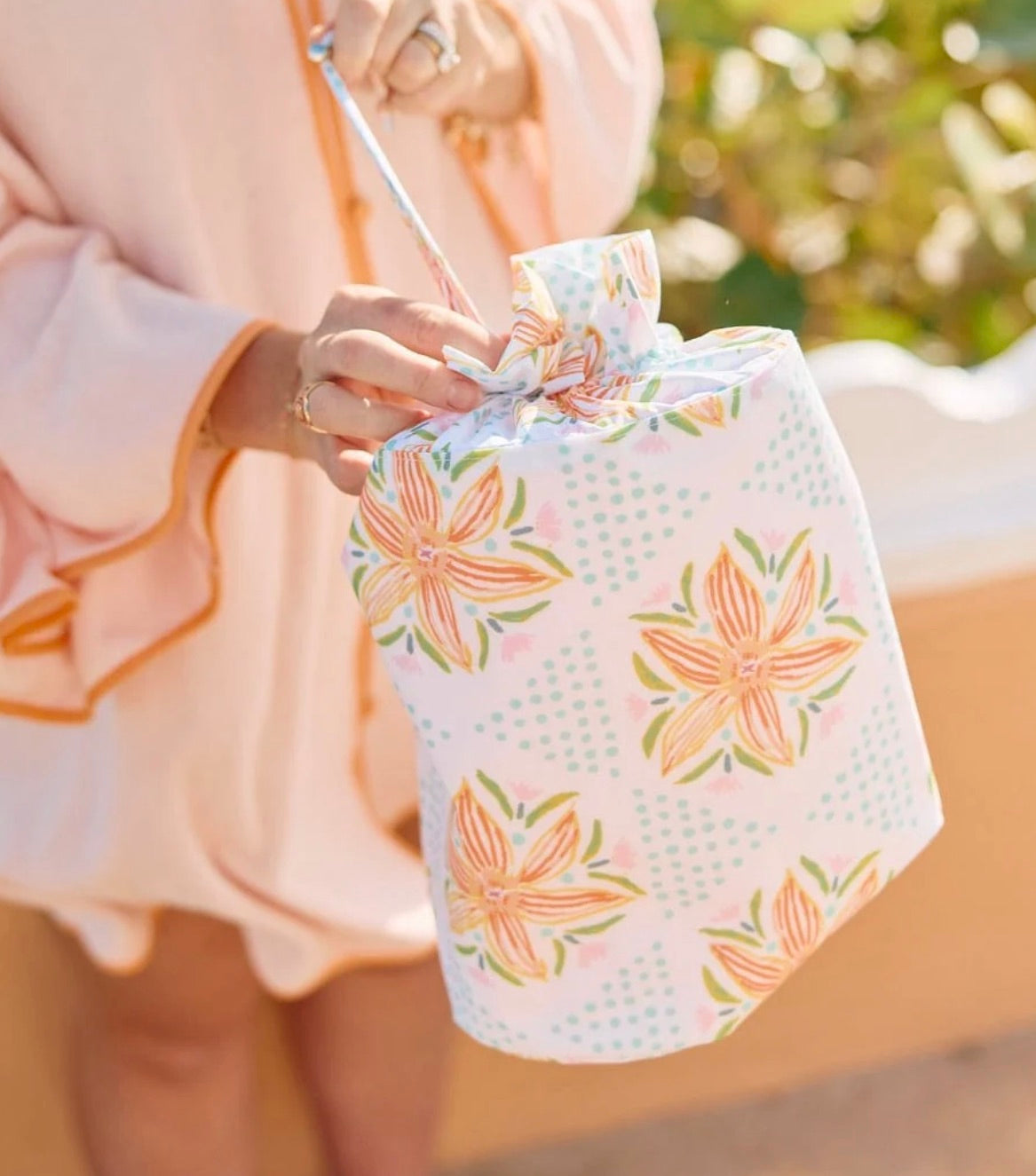 A person in a peach outfit holds the Weezie x Lulie Wallace Wet Bag by Weezie Towels, a floral-patterned, water-resistant bag with a ribbon top—ideal as a wet swimsuit or beach travel bag.