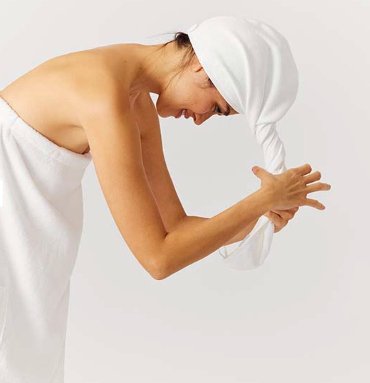 A person leans forward, twisting a Weezie Towels Hair Towel Wrap around their head while wearing a matching white towel.