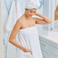A woman stands in a bathroom, smiling and looking down while wearing a white towel and the Weezie Towels Hair Towel Wrap around her hair.