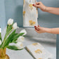 A person hangs the Weezie Towels x Lulie Wallace Louisa Powder Towel with yellow floral patterns on a metal ring above a marble counter, alongside another cotton towel and a vase of white tulips.