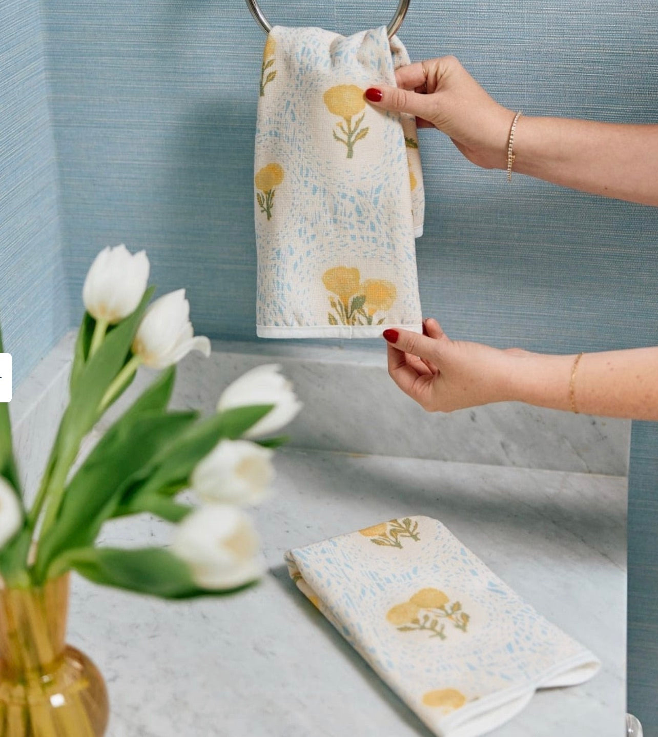 A person hangs the Weezie Towels x Lulie Wallace Louisa Powder Towel with yellow floral patterns on a metal ring above a marble counter, alongside another cotton towel and a vase of white tulips.