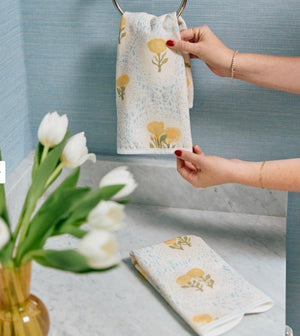 A person hangs the Weezie Towels x Lulie Wallace Louisa Powder Towel with yellow floral patterns on a metal ring above a marble counter, alongside another cotton towel and a vase of white tulips.