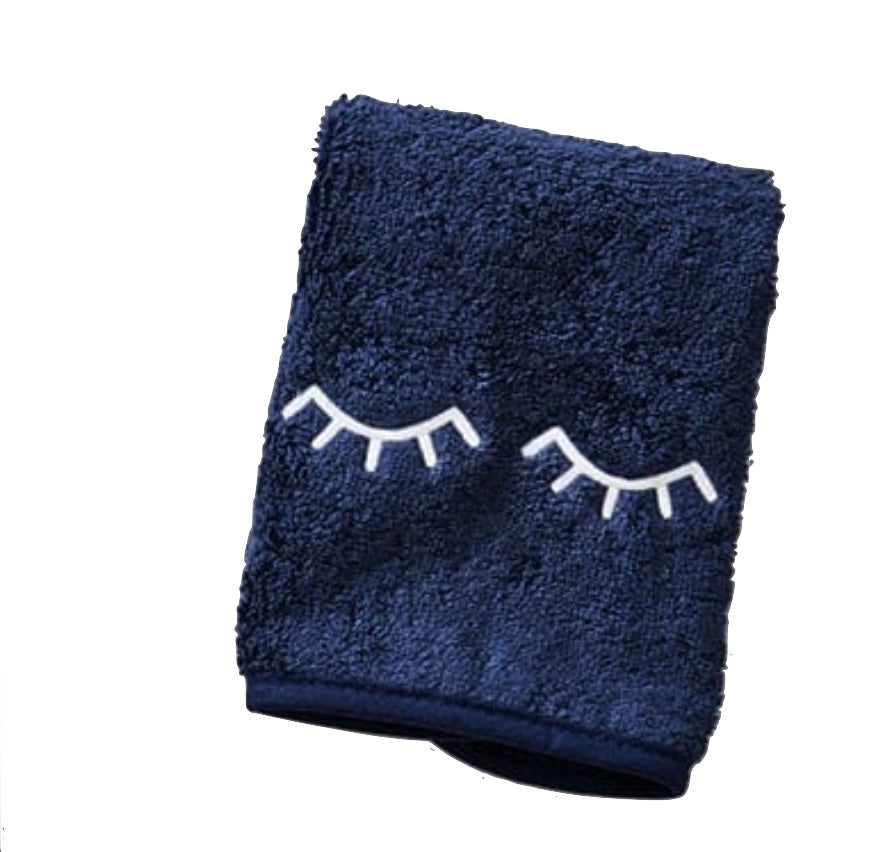 The Weezie Towels Makeup Towel is a navy terry towel with closed eyes and eyelashes embroidered in white, designed to be stain resistant for makeup removal.