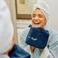 A woman with a towel wrapped around her head smiles in front of a mirror, holding the Weezie Towels Makeup Towel with embroidered closed eyes up to her face.