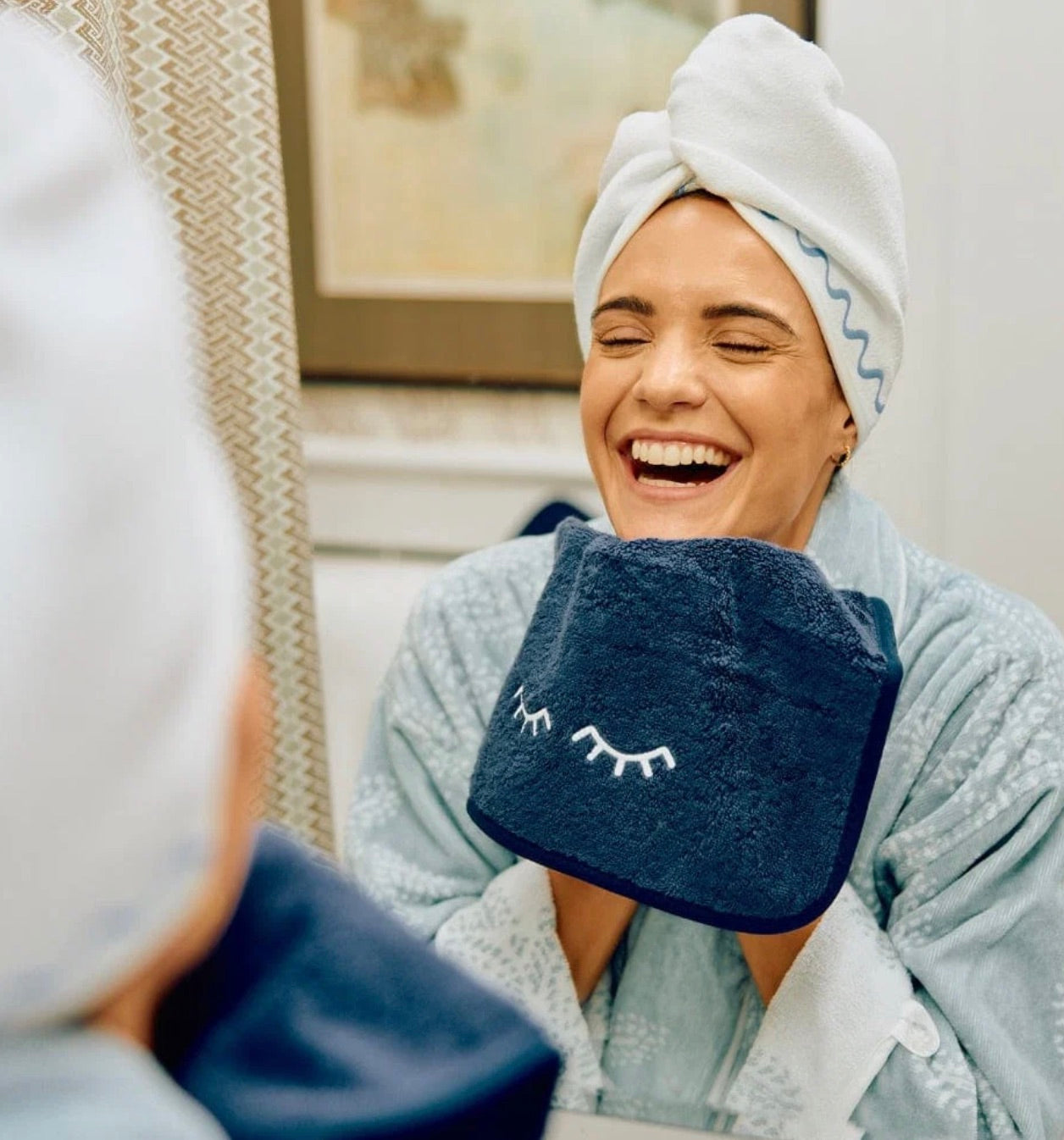 A woman with a towel wrapped around her head smiles in front of a mirror, holding the Weezie Towels Makeup Towel with embroidered closed eyes up to her face.