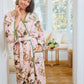 A woman wearing the Vivian Cherry Blossom Robe by Weezie Towels stands smiling in a doorway, with a bright living room and a bouquet of flowers behind her.