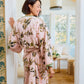 A woman stands in a sunlit room wearing the Vivian Cherry Blossom Robe by Weezie Towels, featuring a plush feel and floral design. She looks over her shoulder, smiling.