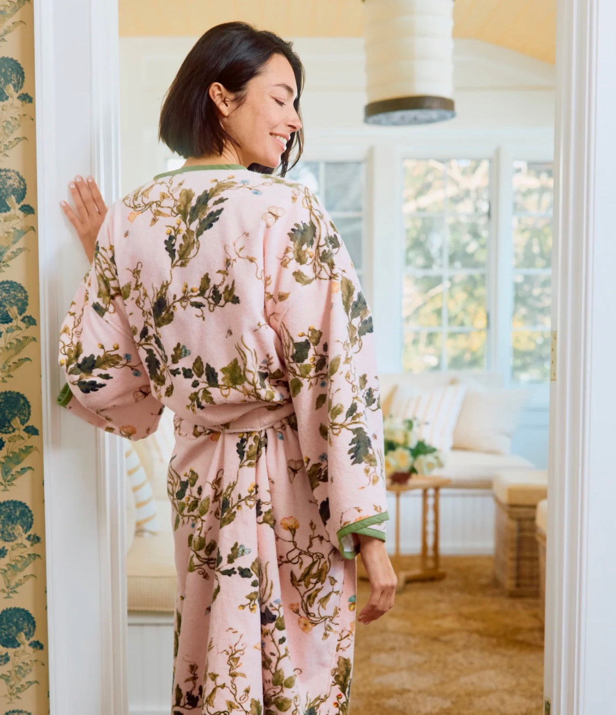 A woman stands in a sunlit room wearing the Vivian Cherry Blossom Robe by Weezie Towels, featuring a plush feel and floral design. She looks over her shoulder, smiling.