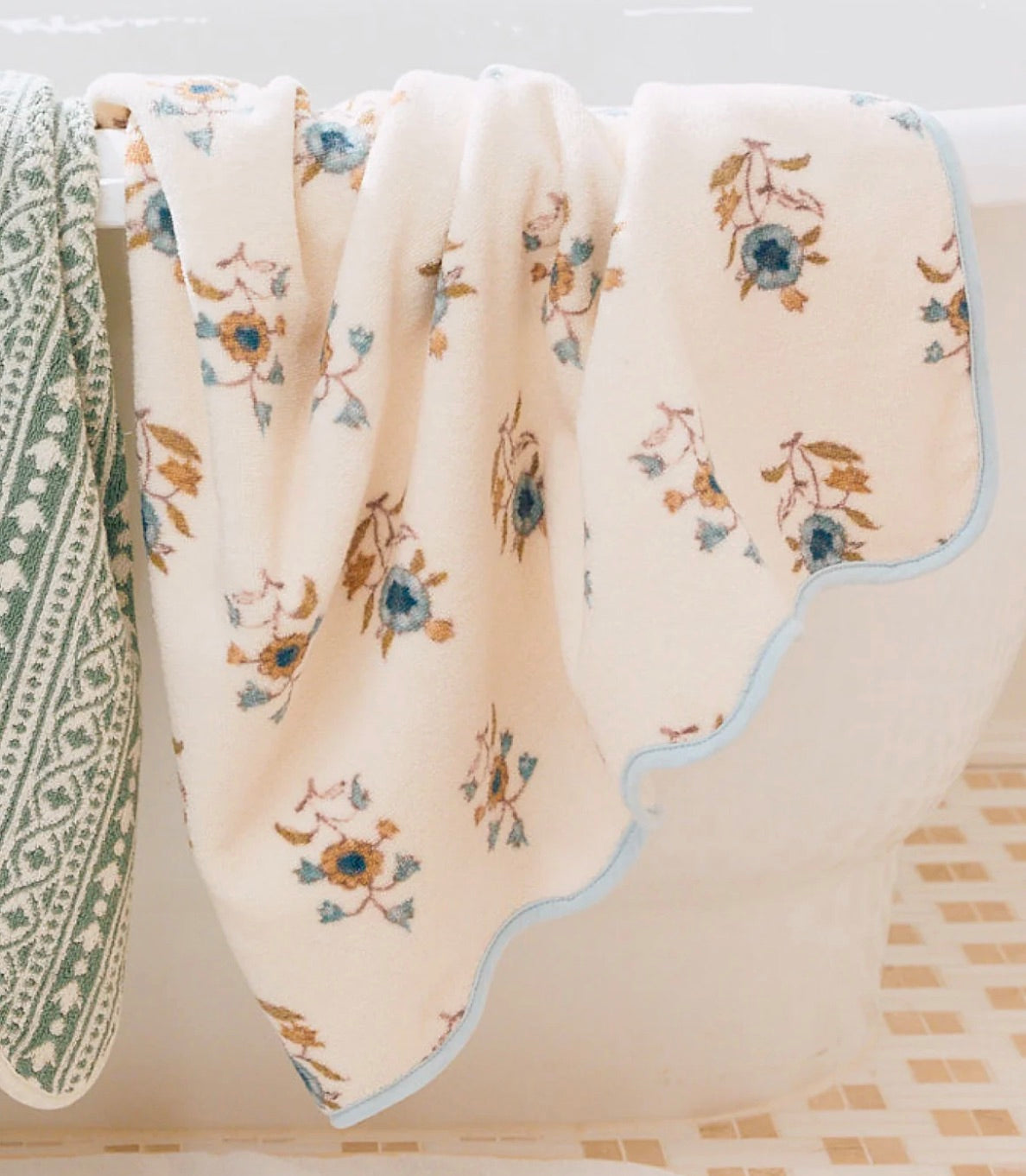 A towel from the Floral Scallop Cora Towel Collection by HIVE Home, Gift and Garden hangs over the bathtub next to a green and white towel, adding a touch of luxury to your bathroom.