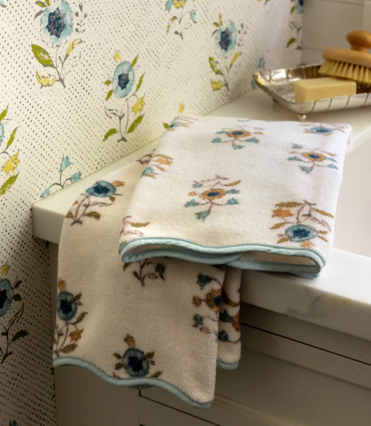 Two folded Floral Scallop Cora Towels from HIVE Home, Gift and Garden rest on a white countertop beside a tray with a brush and soap, bringing a touch of luxury to your bathroom space.