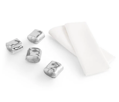 Four polished, hammered-metal napkin rings next to two folded cotton napkins from the Michael Aram Reflective Napkin and Napkin Ring Gift Set.