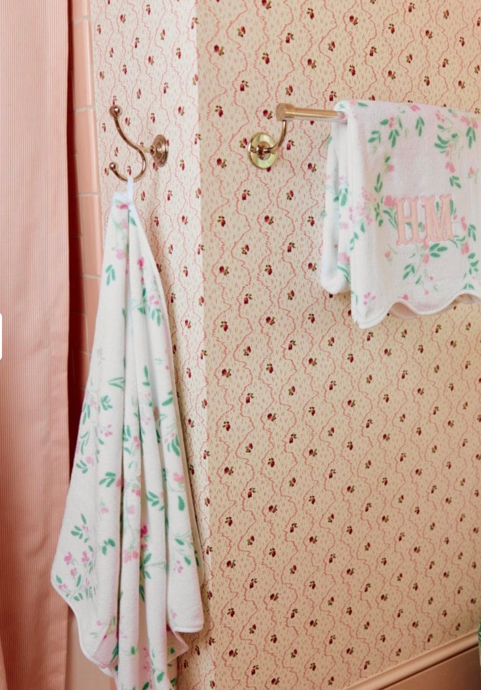 A peach and pink bathroom with floral wallpaper features a Weezie Towels Alice Floral Scallop Towel, hand-finished with scallop trim and absorbent terry on a hook, plus a monogrammed towel on a gold bar.