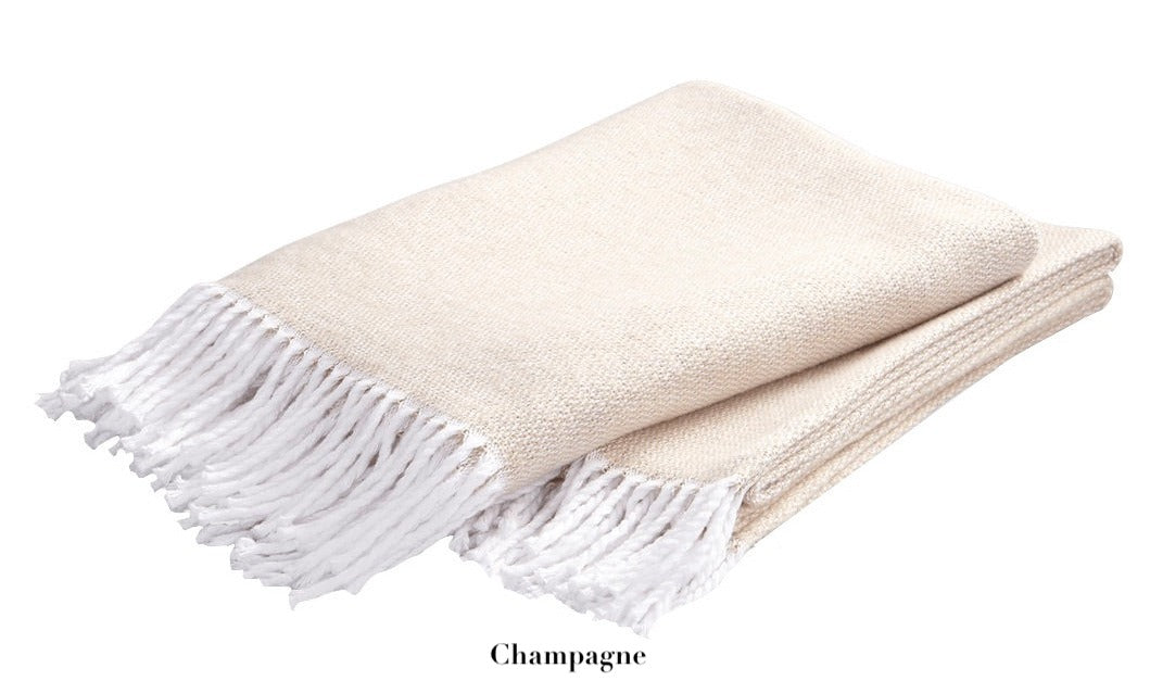 The Pezzo Throw Collection by Matouk features a folded beige "Champagne" throw with white fringed edges, crafted from OEKO-TEX certified brushed cotton.
