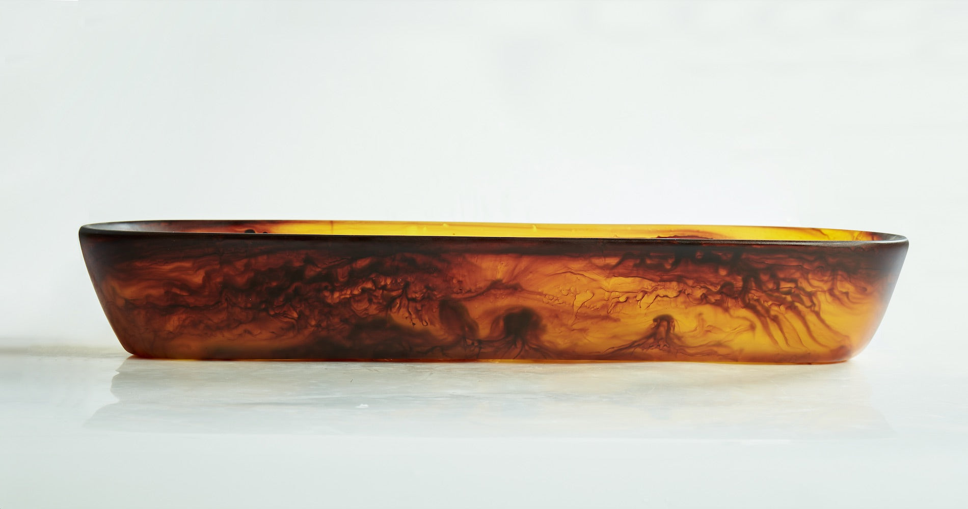 The Tortoise Resin Serveware Collection by Nashi Home features a rectangular amber-hued dish with marbled patterns. Shatter resistant and elegant, it adds warmth to any decor when displayed on a white surface.