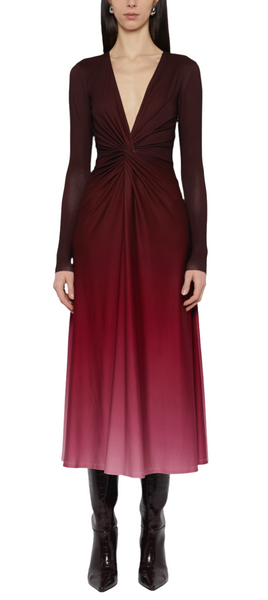 A person wearing a Prabal Gurung Twist Front Long Sleeve Dress with a plunging V-neck in shades of red and pink, paired with dark boots. The ombre design dress features a twisted knot detail at the waist.