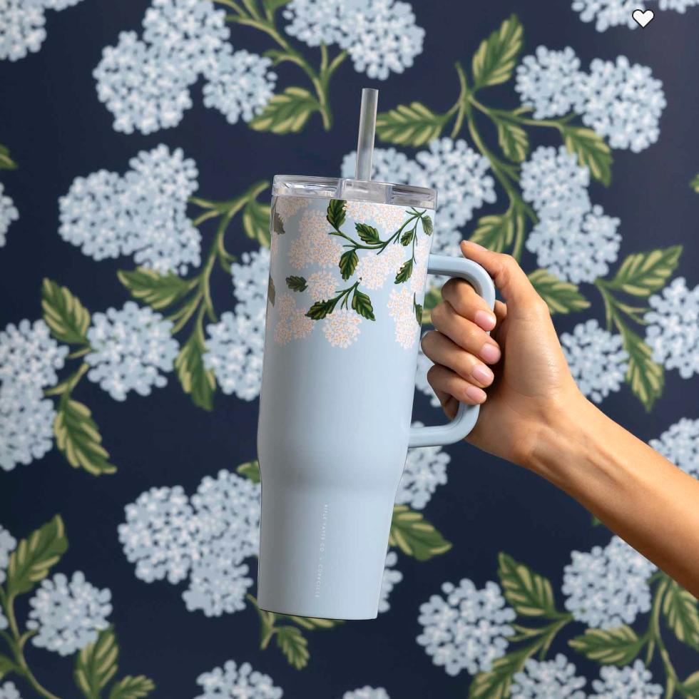 A hand holds the Rifle Paper Co. Blue Hydrangea Cruiser, a large 40-oz straw cup with a floral design and spill-proof lid, in front of matching floral wallpaper.
