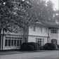 Black and white photo featured on the cover of Houses That We’ve Dreamt Of by Penguin Random House, showcasing a two-story home with expansive windows, manicured bushes, and trees in the background. The gracefully curving driveway highlights the elegance of contemporary American design, inviting readers to imagine refined interiors within.