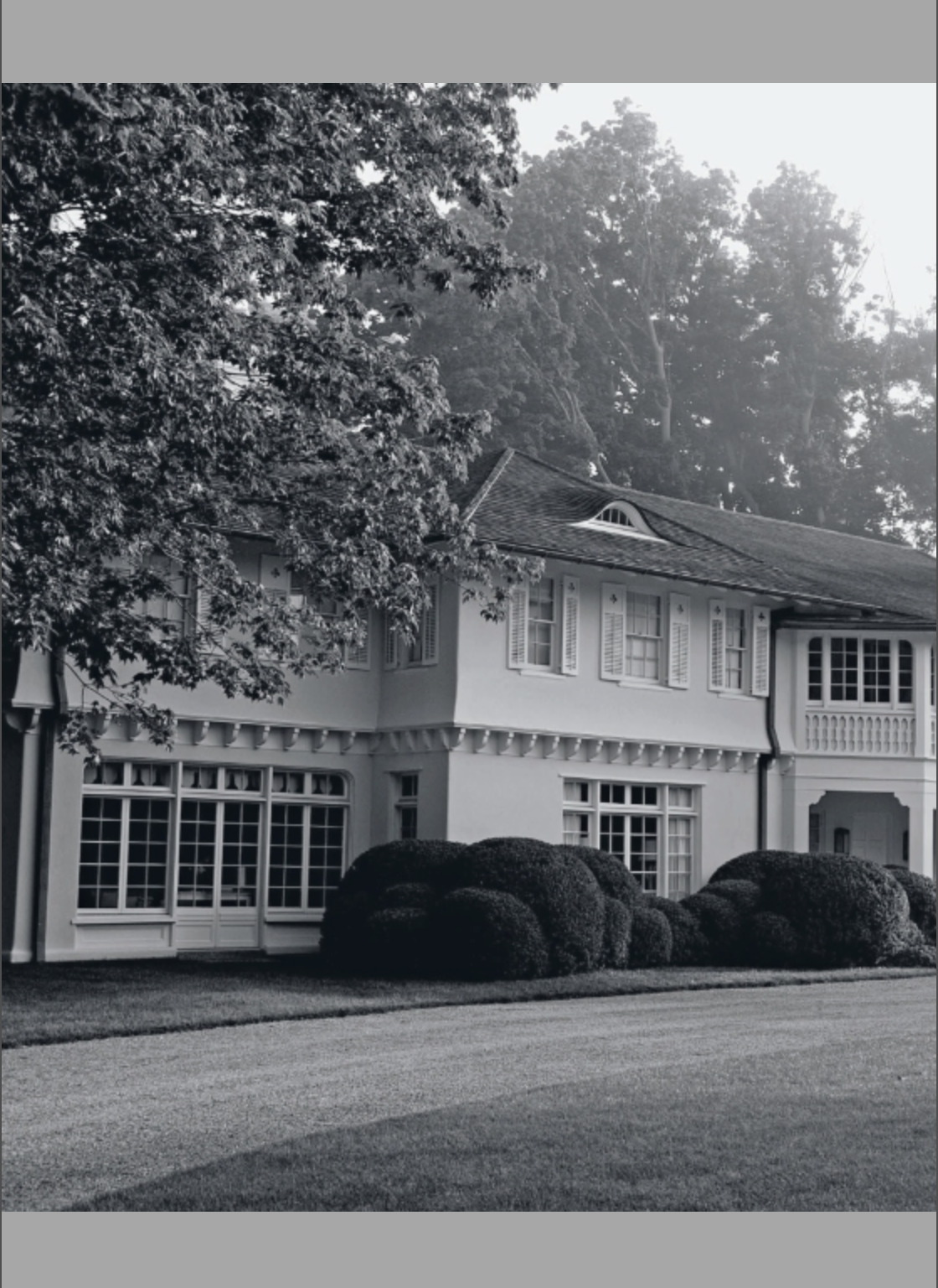 Black and white photo featured on the cover of Houses That We’ve Dreamt Of by Penguin Random House, showcasing a two-story home with expansive windows, manicured bushes, and trees in the background. The gracefully curving driveway highlights the elegance of contemporary American design, inviting readers to imagine refined interiors within.