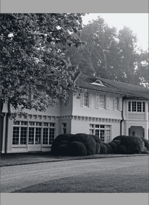 Black and white photo featured on the cover of Houses That We’ve Dreamt Of by Penguin Random House, showcasing a two-story home with expansive windows, manicured bushes, and trees in the background. The gracefully curving driveway highlights the elegance of contemporary American design, inviting readers to imagine refined interiors within.