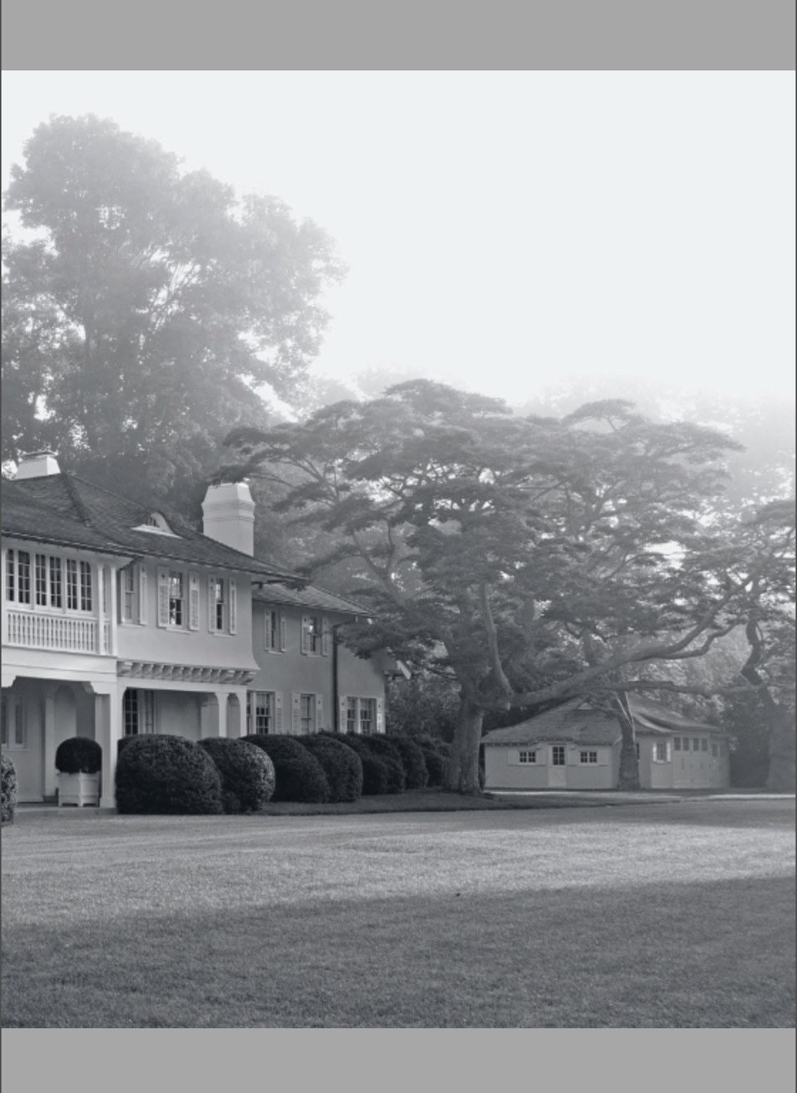 Black and white photo from “Houses That We’ve Dreamt Of” by Penguin Random House, depicting a large house with a porch, trimmed hedges, and an expansive lawn—its graceful architecture hinting at interiors where period and contemporary design coexist in harmony.