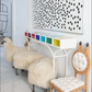A row of furry stools, a wooden chair with white cushions, a colorful cubby shelf, and *Houses That We’ve Dreamt Of* by Penguin Random House displayed above reflect the playful sophistication found in contemporary American design.