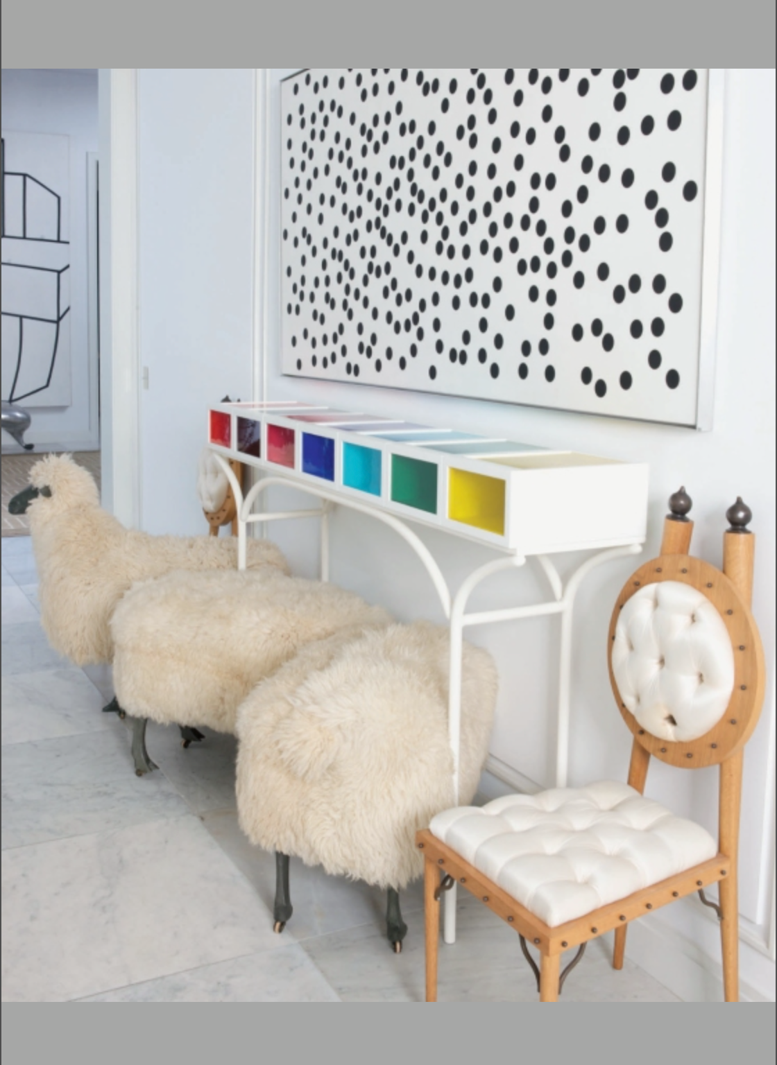 A row of furry stools, a wooden chair with white cushions, a colorful cubby shelf, and *Houses That We’ve Dreamt Of* by Penguin Random House displayed above reflect the playful sophistication found in contemporary American design.