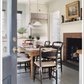 The bright kitchen featured in “The Great American House” by Rizzoli showcases a wooden table, four chairs, hanging lights, a fireplace, and potted plants on the windowsill beside a stainless steel refrigerator—an inviting space that beautifully captures the charm of traditional houses through thoughtful design elements.