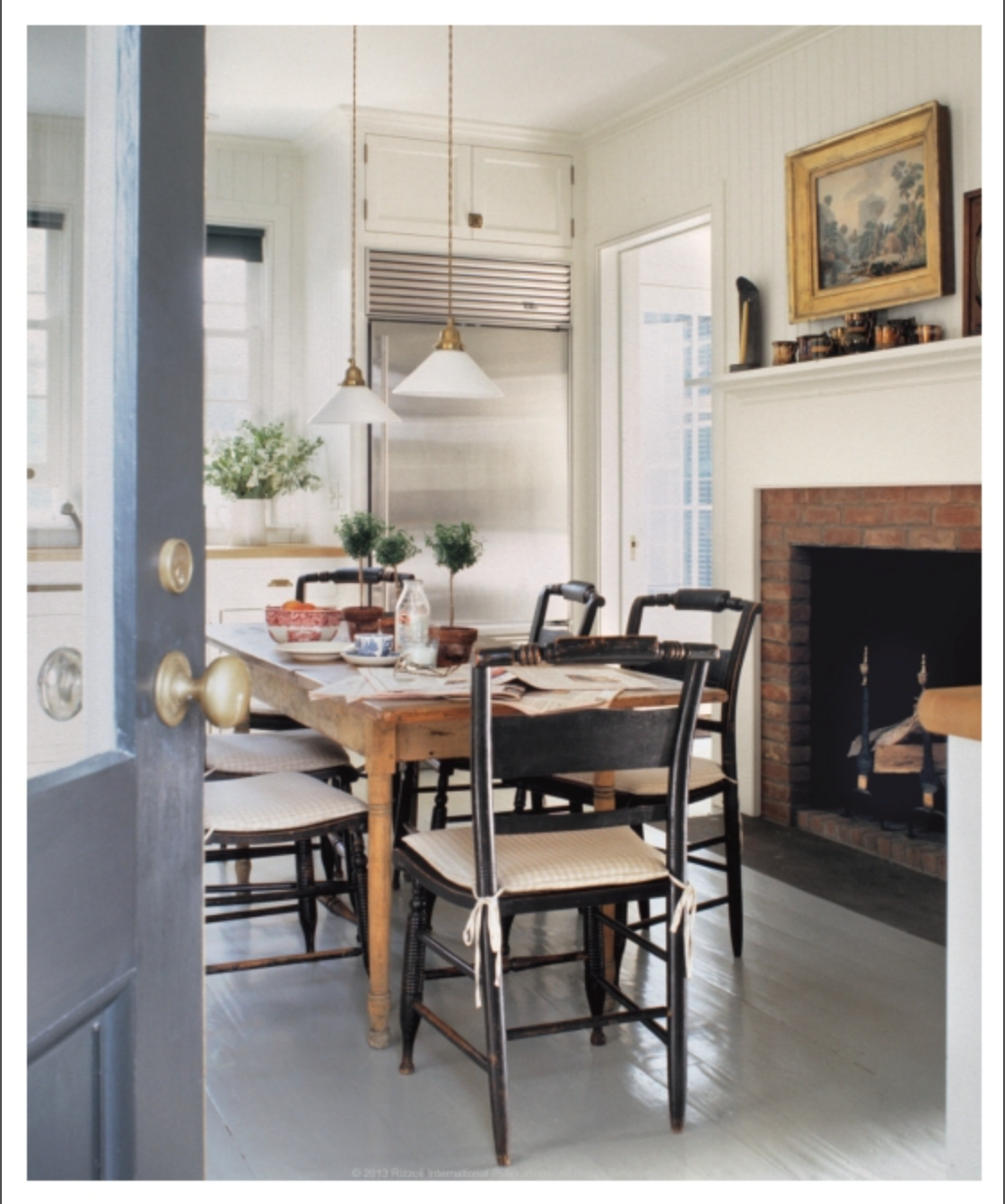 The bright kitchen featured in “The Great American House” by Rizzoli showcases a wooden table, four chairs, hanging lights, a fireplace, and potted plants on the windowsill beside a stainless steel refrigerator—an inviting space that beautifully captures the charm of traditional houses through thoughtful design elements.