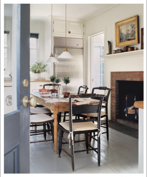 The bright kitchen featured in “The Great American House” by Rizzoli showcases a wooden table, four chairs, hanging lights, a fireplace, and potted plants on the windowsill beside a stainless steel refrigerator—an inviting space that beautifully captures the charm of traditional houses through thoughtful design elements.