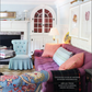 A living room featuring a purple tufted sofa, floral armchair, blue chair, built-in cabinets, and various pillows embodies thoughtful interior design. A lamp and artwork are visible, with a copy of Rizzoli’s *Gardens of Style* open on the sofa.