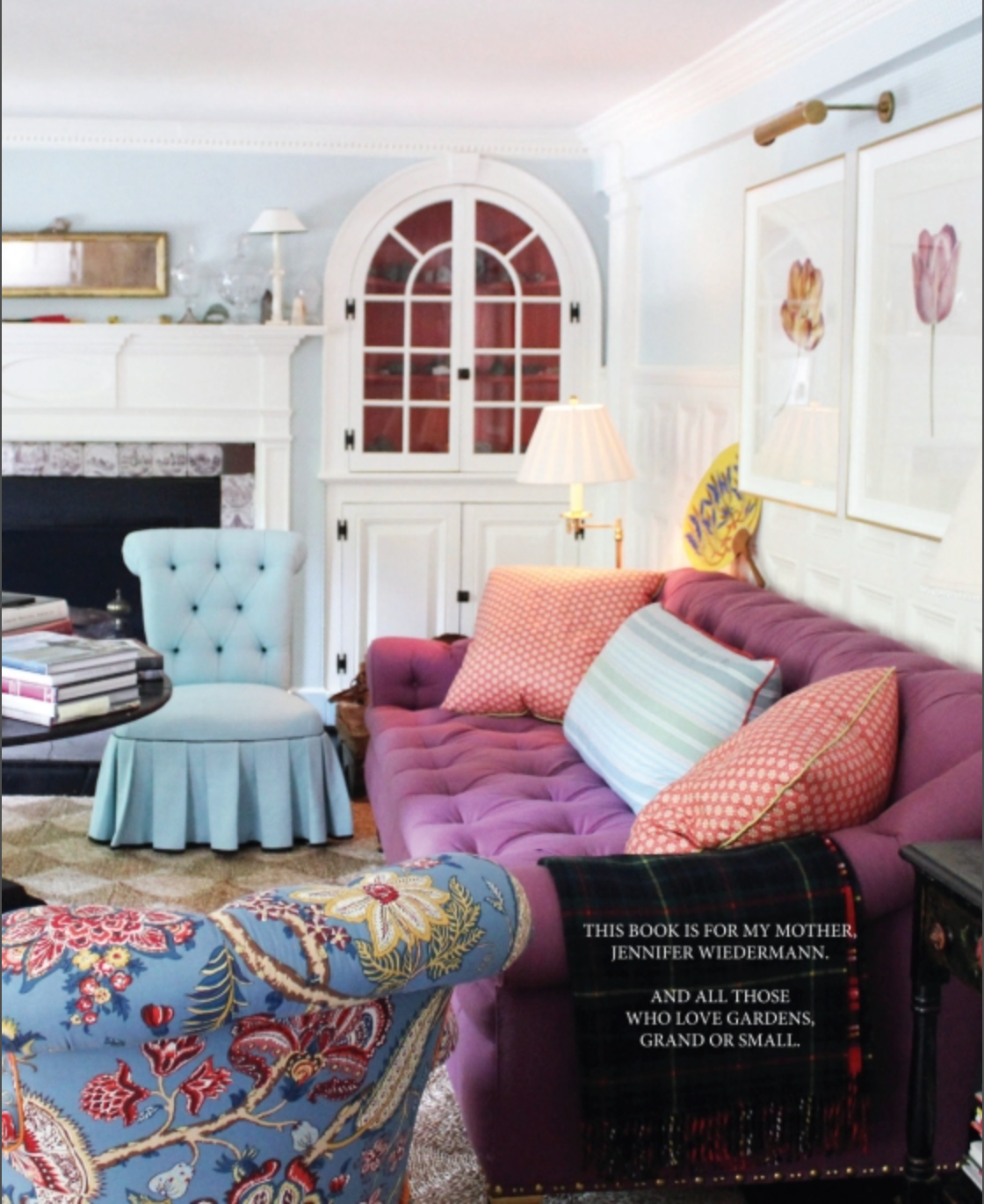 A living room featuring a purple tufted sofa, floral armchair, blue chair, built-in cabinets, and various pillows embodies thoughtful interior design. A lamp and artwork are visible, with a copy of Rizzoli’s *Gardens of Style* open on the sofa.