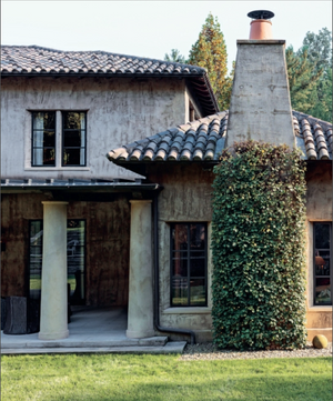 Two-story stucco house with tile roof, covered porch supported by columns, tall chimney draped in climbing ivy, and a green lawn in front—a classic example of Southern homes reminiscent of those featured in *Creating Home: Design for Living* by Rizzoli.