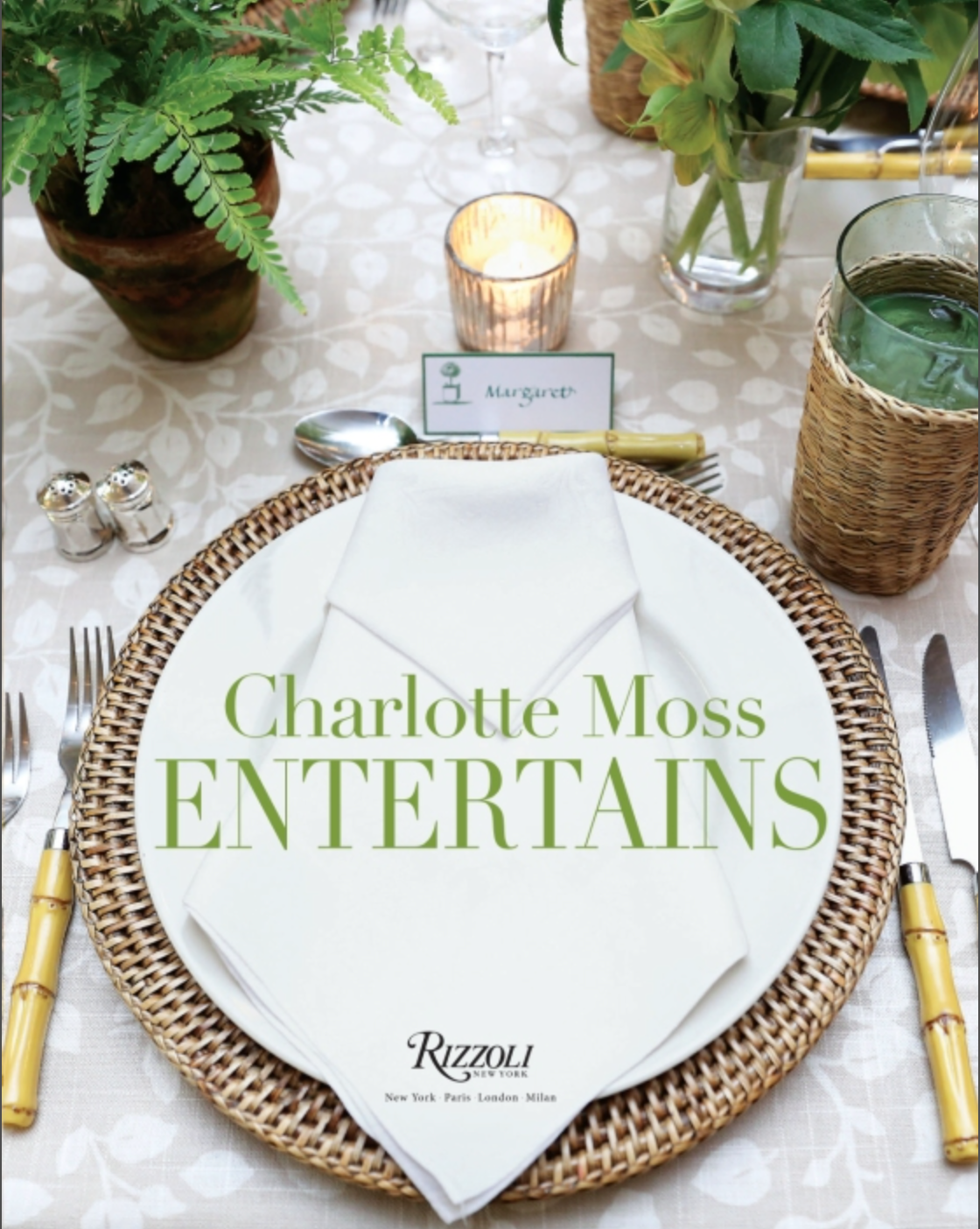 A place setting with a woven charger, crisp white napkin, and refined cutlery captures the spirit of gracious hospitality. At each seat, the Charlotte Moss Entertains plate by Rizzoli sets an elegant tone. Lush greenery, a softly glowing candle, and personalized name cards complete the inviting tablescape.