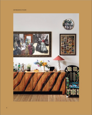 Featuring “Beata Heuman, Every Room Should Sing” by Rizzoli, this space showcases a wooden sideboard with a zebra pattern adorned with flowers and a lamp, infusing the room with Scandinavian flair; vibrant art and photos hang above, beside an upholstered floral chair thoughtfully arranged by an interior designer.
