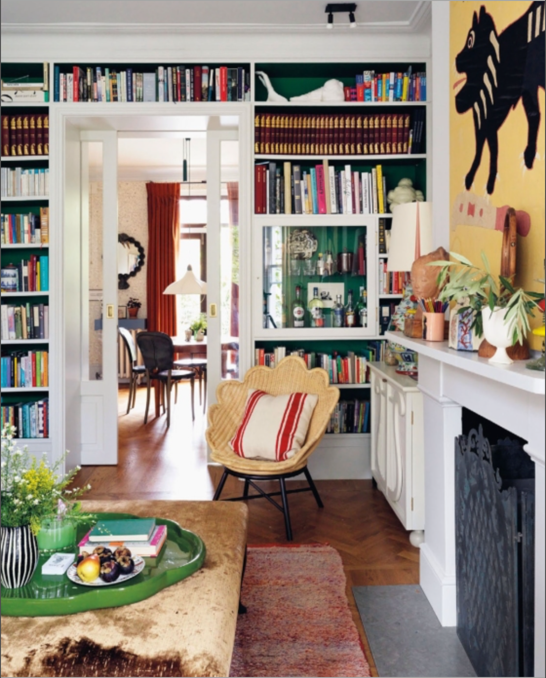A cozy living room with bookshelves, a chair, a fireplace, and a green tray atop a velvet sofa featuring bespoke furnishings from Beata Heuman’s Every Room Should Sing (Rizzoli); the subtly Scandinavian-styled dining room is seen through glass doors.