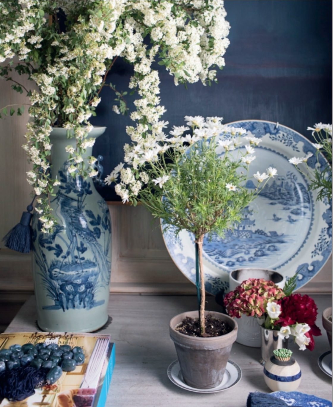 Elevate your home decor with a stylish tabletop display featuring A Home For All Seasons by Penguin Random House, paired with a large blue-and-white vase of white flowers, a small potted plant, a plate, assorted flowers in jars, and curated accents—perfect for entertaining in style.