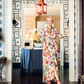 A woman in a floral dress stands in a doorway of a room adorned with patterned wallpaper, a table displaying orchids, and a red hanging light fixture, embodying the spirit of elegant home decor showcased in “A Home For All Seasons” by Penguin Random House.