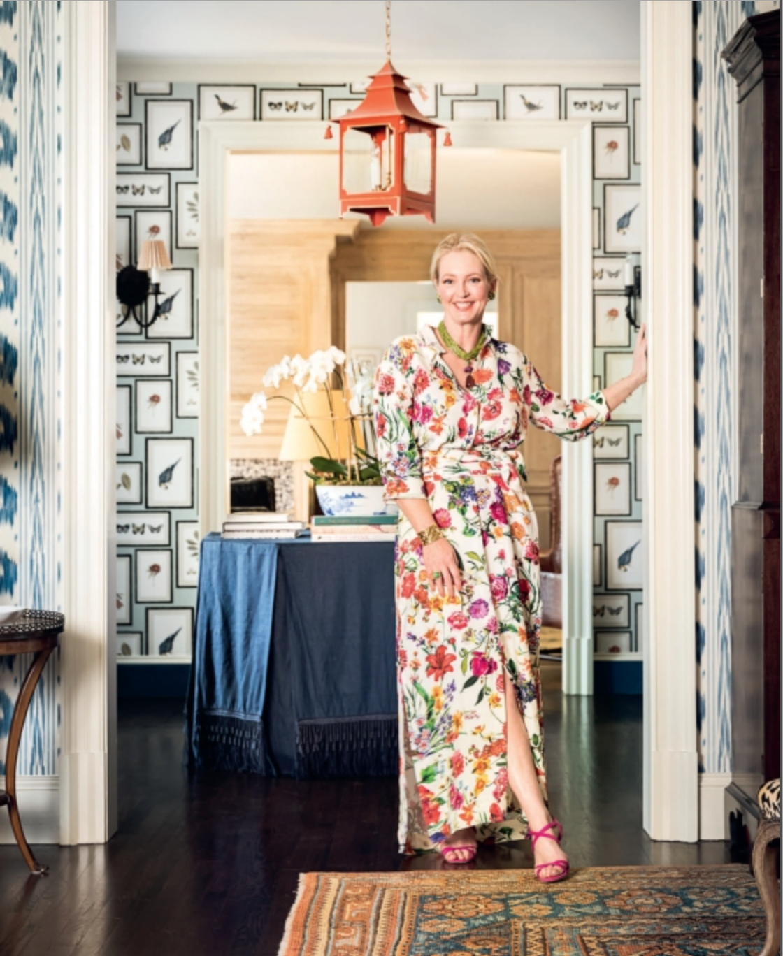 A woman in a floral dress stands in a doorway of a room adorned with patterned wallpaper, a table displaying orchids, and a red hanging light fixture, embodying the spirit of elegant home decor showcased in “A Home For All Seasons” by Penguin Random House.