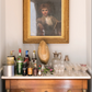 Beneath a framed portrait in a gold frame rests a wooden sideboard adorned with assorted liquor bottles, glassware, sculpted accents, and an ice bucket—an arrangement evocative of the dreamy tablescapes and personal anecdotes found in An Entertaining Story by Rizzoli.