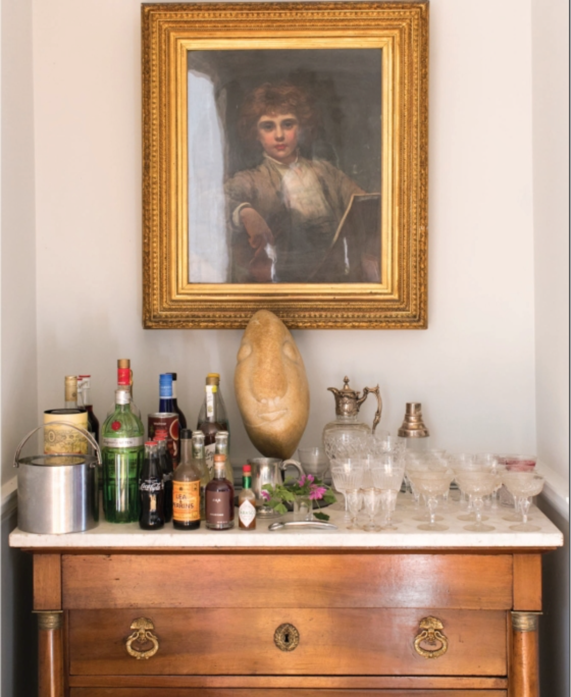 Beneath a framed portrait in a gold frame rests a wooden sideboard adorned with assorted liquor bottles, glassware, sculpted accents, and an ice bucket—an arrangement evocative of the dreamy tablescapes and personal anecdotes found in An Entertaining Story by Rizzoli.