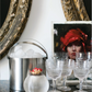 A metal ice bucket, a glass bowl of matches, five crystal glasses, and a portrait painting are arranged on a marble surface, evoking the dreamy tablescapes described in *An Entertaining Story* by Rizzoli—perfect for sharing family anecdotes and sparkling drinks.