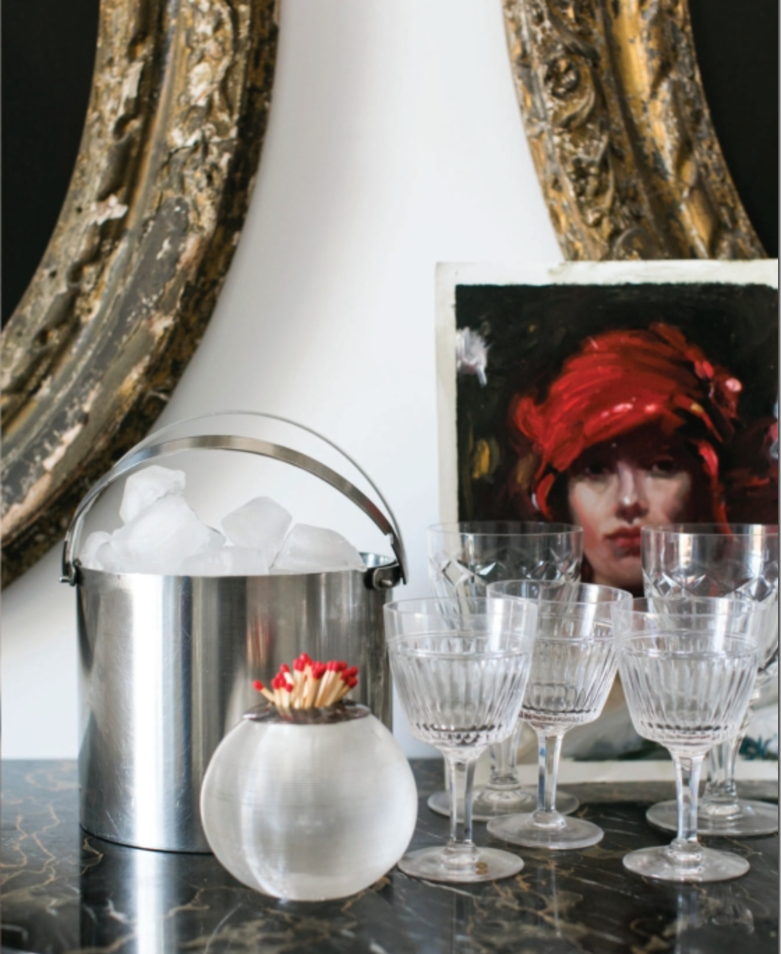 A metal ice bucket, a glass bowl of matches, five crystal glasses, and a portrait painting are arranged on a marble surface, evoking the dreamy tablescapes described in *An Entertaining Story* by Rizzoli—perfect for sharing family anecdotes and sparkling drinks.
